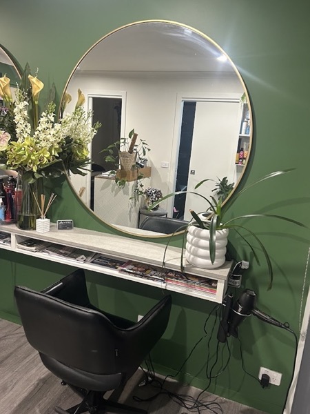 A green wall with a round mirror above a shelf holding flowers